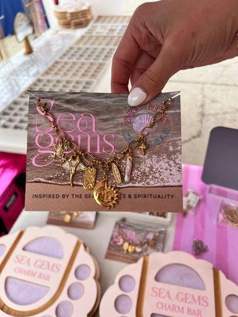 Close-up of a gold ocean-inspired charm bracelet with sun, shell and surfboard charms on a printed display card, held by a hand with white nail polish over a pastel market jewelry display