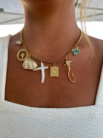 Close-up of a beachy gold charm necklace on a sun-kissed neckline, chunky chain with compass medallion, striped shell, baroque pearl cross, engraved 'D' tag, turquoise clover and gold fish-hook charm over a white tank top.