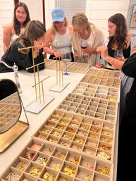 Group of women at a home jewelry party browsing organized trays of gold charms, earrings and findings on a kitchen island with display stands and wine glasses.