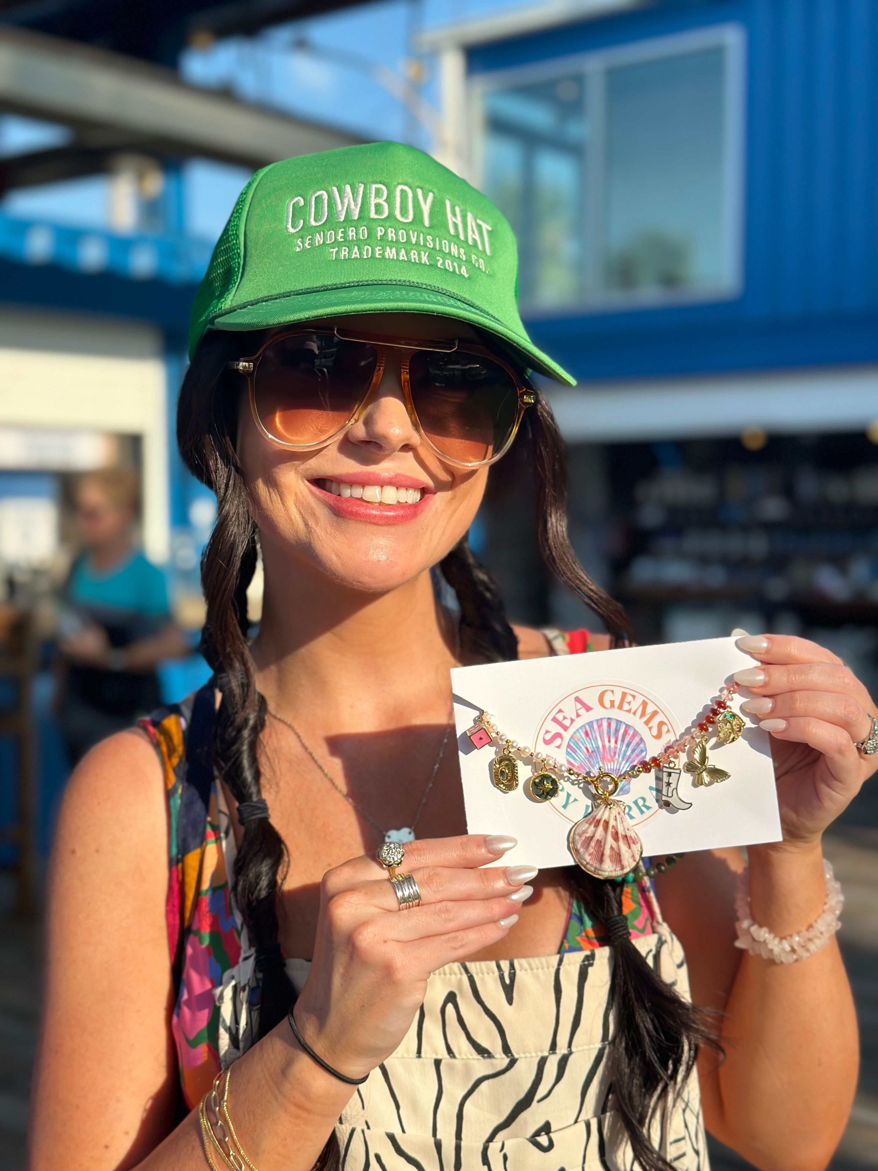 Smiling woman in a green cap and aviator sunglasses at a sunny seaside market holding a seashell charm necklace on a display card — boho beach jewelry and summer style.