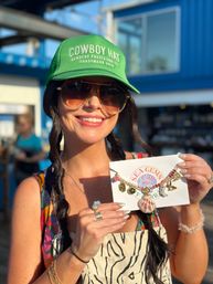 Smiling woman in a green cap and aviator sunglasses at a sunny seaside market holding a seashell charm necklace on a display card — boho beach jewelry and summer style.