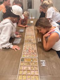Group of women leaning over a long wooden table at a home craft party, selecting gold letter charms, colorful beads and tiny pendants from organized compartment trays for DIY jewelry making.