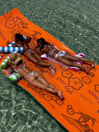 Three friends in colorful bikinis sunbathing on a large orange floating mat in crystal-clear tropical water.