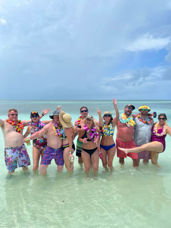 Smiling adults in colorful leis and swimwear posing waist-deep in clear turquoise shallow water on a tropical beach under a bright, cloudy sky