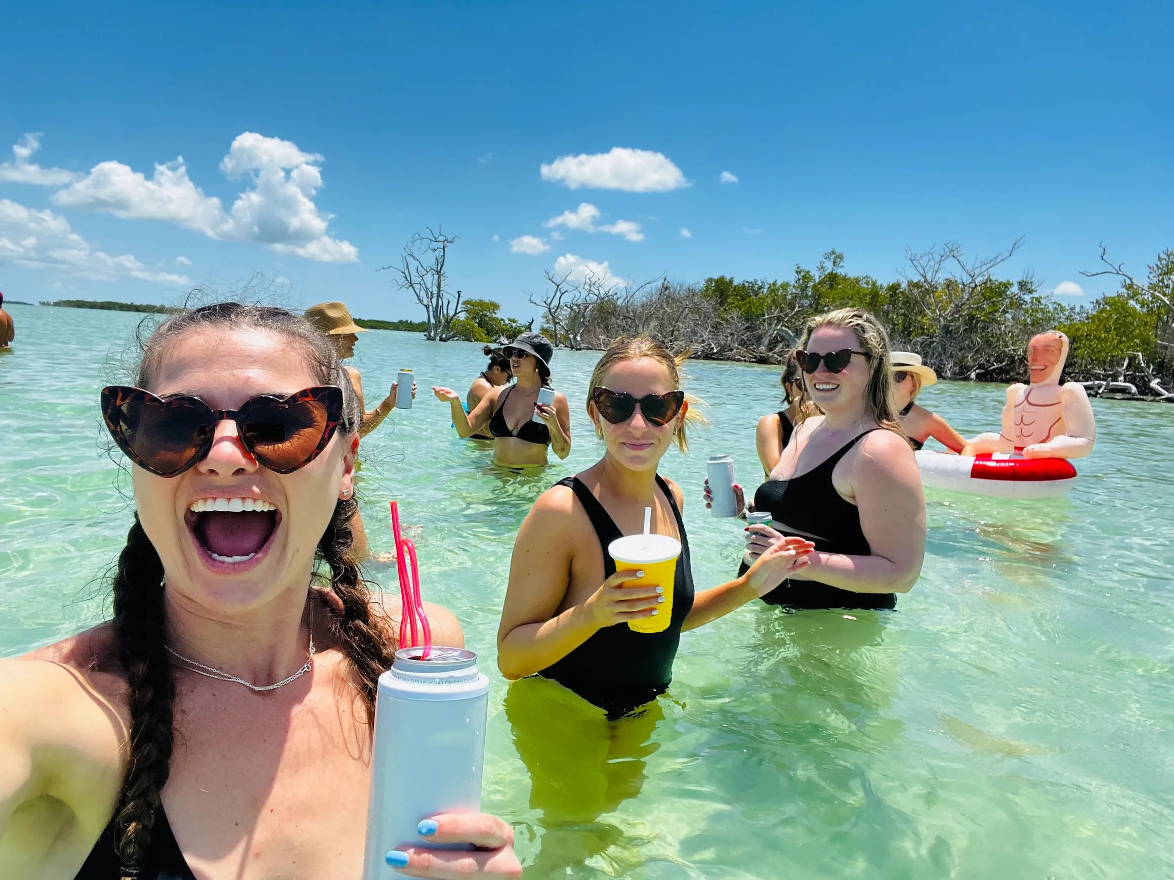Friends in swimsuits laughing and holding drinks while wading in shallow turquoise lagoon near mangroves under a sunny blue sky