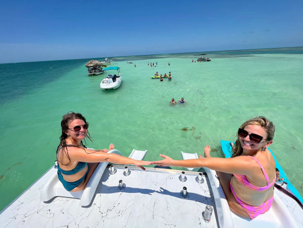 Two women in swimsuits and sunglasses sit on a boat swim platform, reaching hands toward each other over a shallow turquoise sandbar with small boats, waders, and a thatched floating palapa in clear tropical water under a bright blue sky.