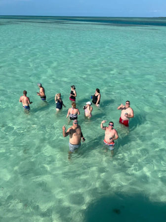 Aerial view of a group of people wading and cheering with drinks in shallow turquoise tropical water under a clear blue sky.