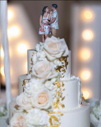 Three-tier white fondant wedding cake with cascading blush roses and gold leaf accents, couple figurine topper, warm bokeh reception lights