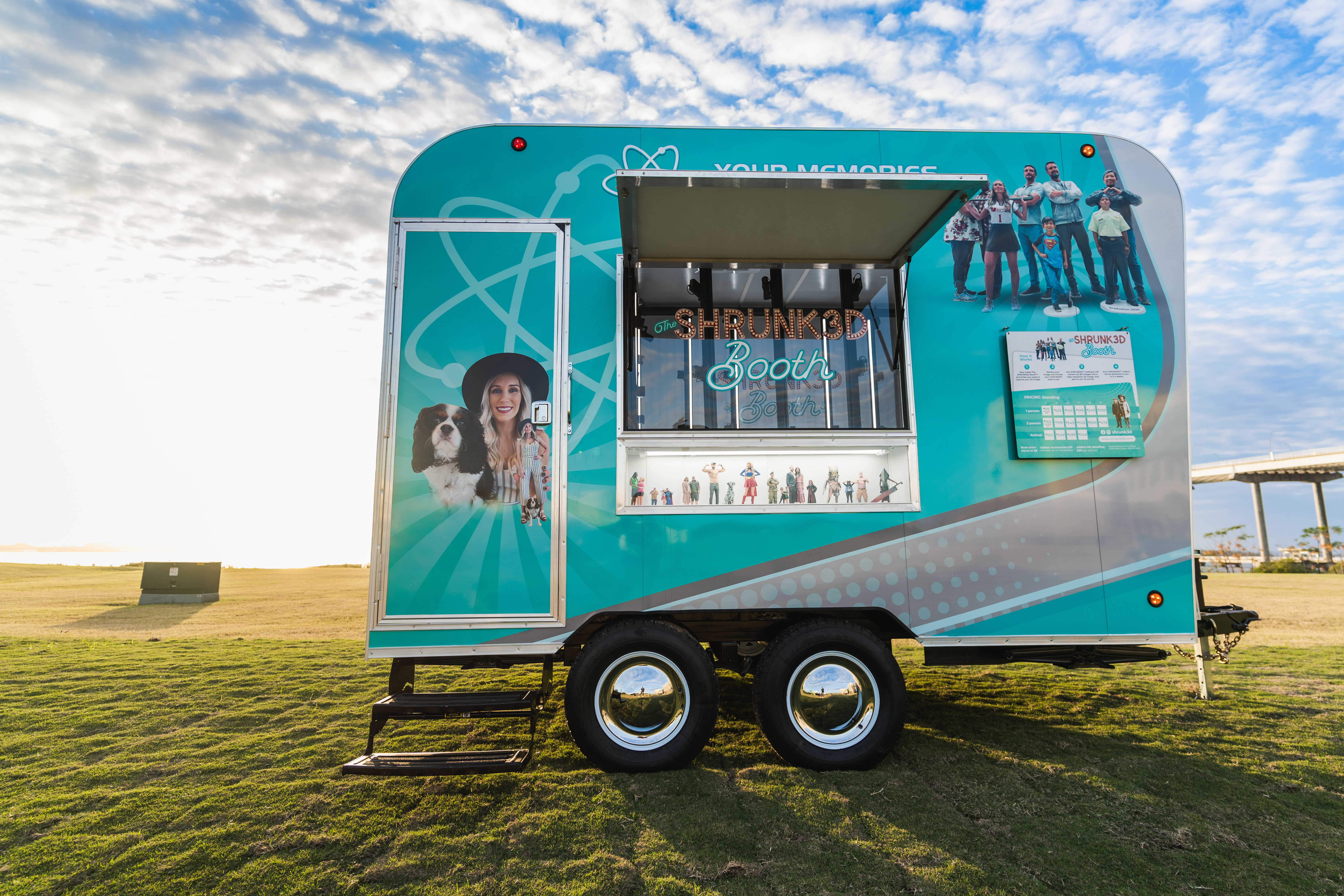 Teal mobile 3D photo-booth trailer parked on a waterfront grassy field under a cloudy sky, open service window displaying miniature figurines and colorful people-and-dog graphics on the side for an outdoor event.