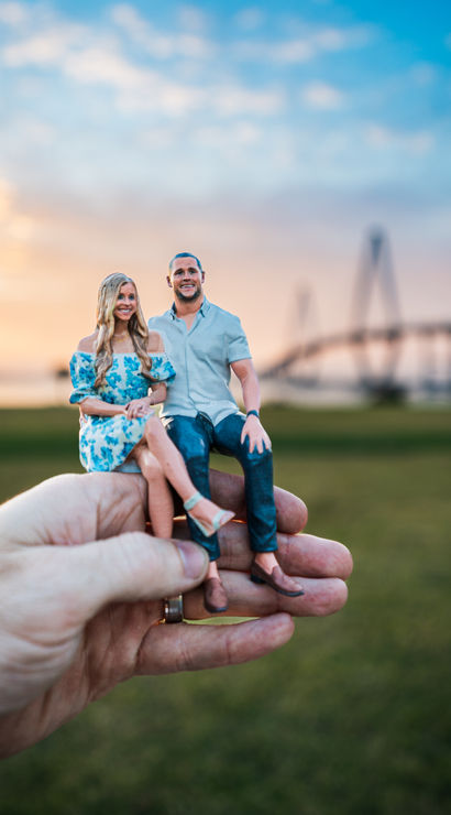 Hand holding tiny 3D-printed couple figurines seated together at sunset in a grassy riverside park with a blurred cable-stayed bridge in the background.