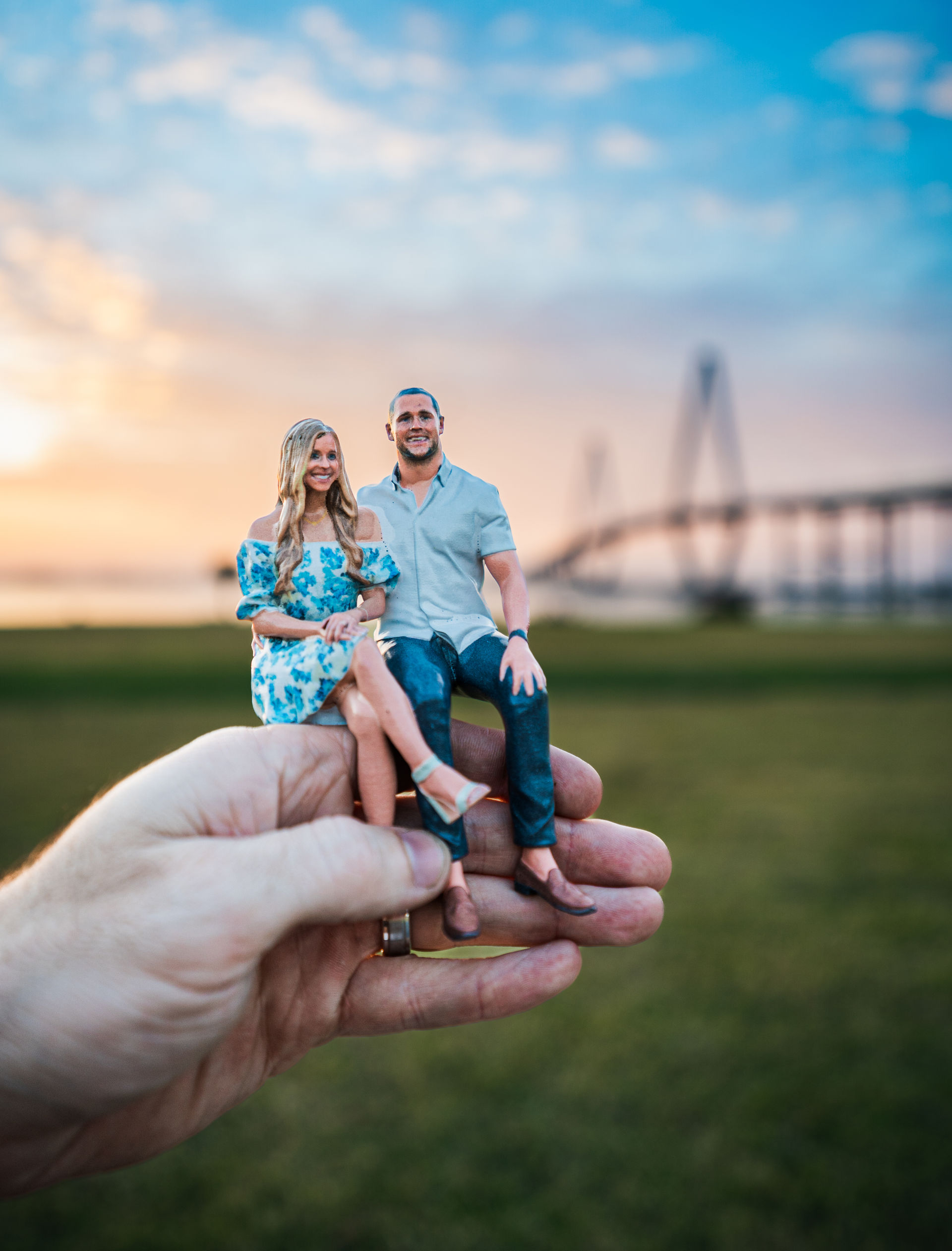 Hand holding tiny 3D-printed couple figurines seated together at sunset in a grassy riverside park with a blurred cable-stayed bridge in the background.