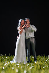 Miniature bride and groom figurines on green grass, smiling and forming a heart with their hands against a black background.