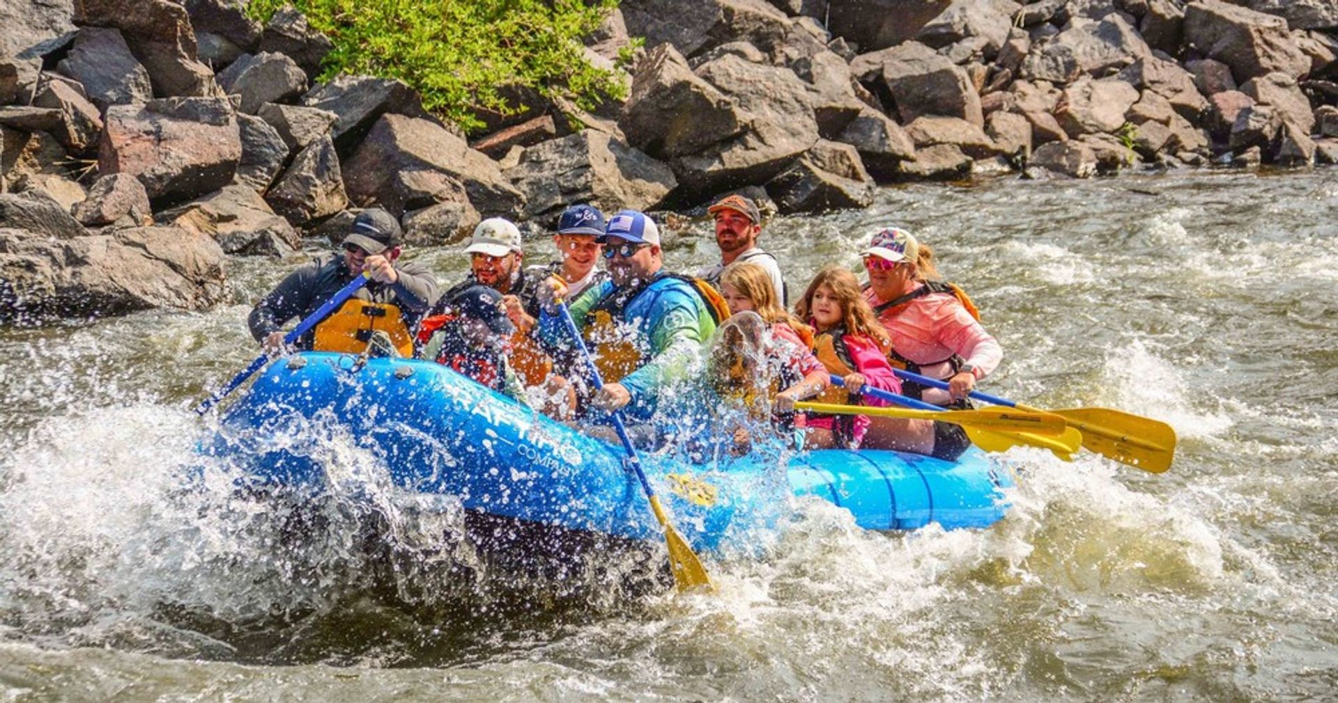 Thrilling whitewater rafting on a blue raft through rocky river rapids, a mixed group paddling and splashing on a sunny mountain river