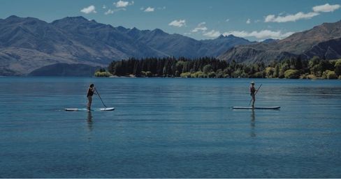 Two stand-up paddleboarders gliding across a calm alpine lake with a forested shoreline and rugged mountains under a bright blue sky.