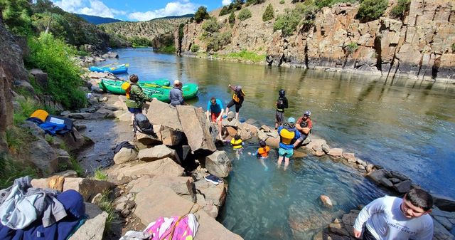 Group of people with colorful inflatable rafts on a rocky riverbank in a scenic canyon; kids in life jackets wading in a small rock-enclosed pool while others prep rafts under a sunny blue sky and steep cliff walls.