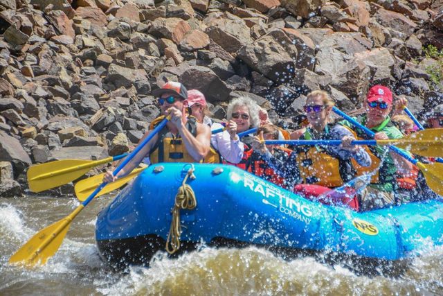 Whitewater rafting group in a bright blue inflatable raft with yellow paddles navigating splashing rapids along a rocky riverbank, passengers wearing life jackets and sunglasses.