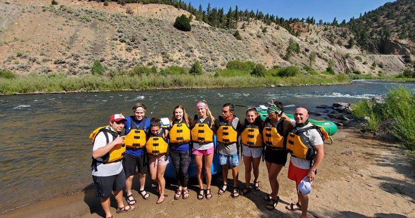 Smiling group of nine rafters in yellow life jackets on a sandy riverbank beside inflatable rafts, ready for a sunny whitewater rafting adventure in a mountain canyon