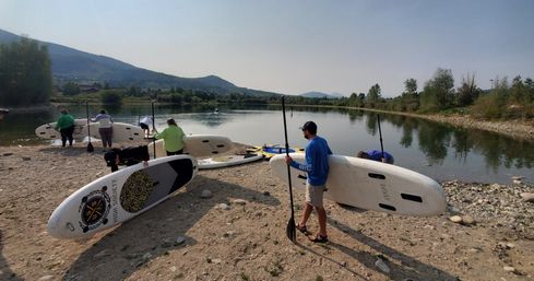 Group gearing up with stand-up paddleboards and paddles on a pebbled river shore, calm reflective water, tree-lined banks and rolling mountains under a clear sky
