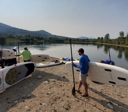 Group gearing up with stand-up paddleboards and paddles on a pebbled river shore, calm reflective water, tree-lined banks and rolling mountains under a clear sky