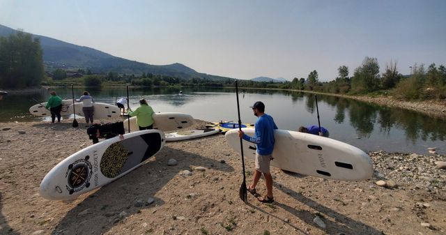 Group gearing up with stand-up paddleboards and paddles on a pebbled river shore, calm reflective water, tree-lined banks and rolling mountains under a clear sky