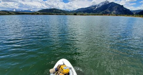 Front of a paddleboard with a yellow dry bag on a calm alpine lake, rippling green water, blue sky and snow-capped mountains on the horizon.