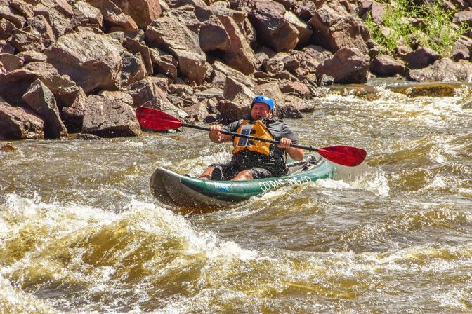Whitewater kayaker navigating rocky mountain river rapids in a green kayak, paddling with red blades while wearing a blue helmet and yellow life jacket.