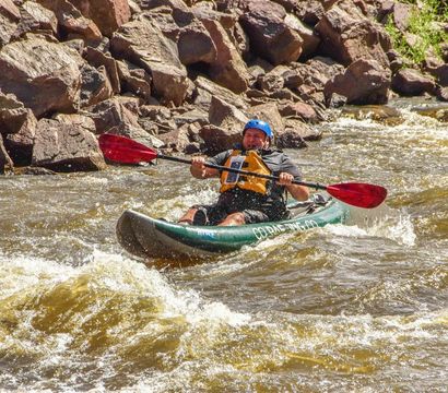 Whitewater kayaker navigating rocky mountain river rapids in a green kayak, paddling with red blades while wearing a blue helmet and yellow life jacket.