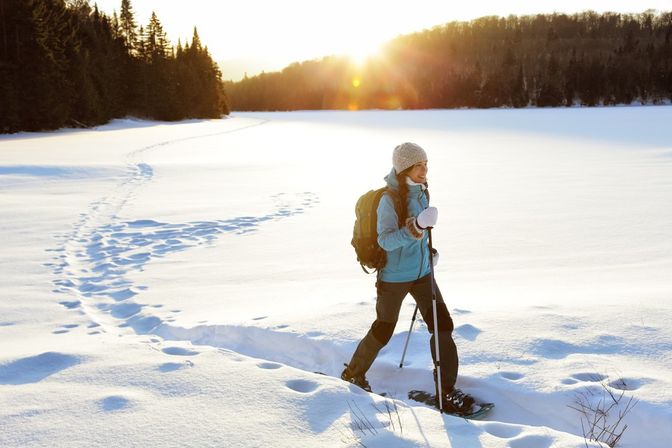 Person snowshoeing across a sunlit snow-covered field at sunrise, wearing a blue jacket, knit hat and backpack, using trekking poles and leaving a winding trail of footprints toward an evergreen tree line.