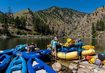 Group prepping colorful inflatable rafts and yellow dry bags on a calm mountain riverbank with pine-covered canyon slopes and a bright blue summer sky — river rafting adventure.