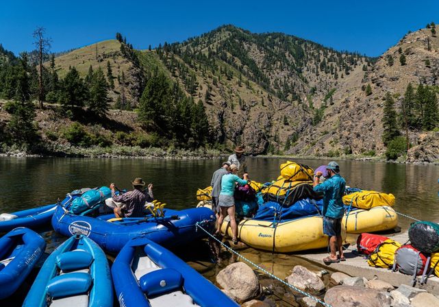 Group prepping colorful inflatable rafts and yellow dry bags on a calm mountain riverbank with pine-covered canyon slopes and a bright blue summer sky — river rafting adventure.