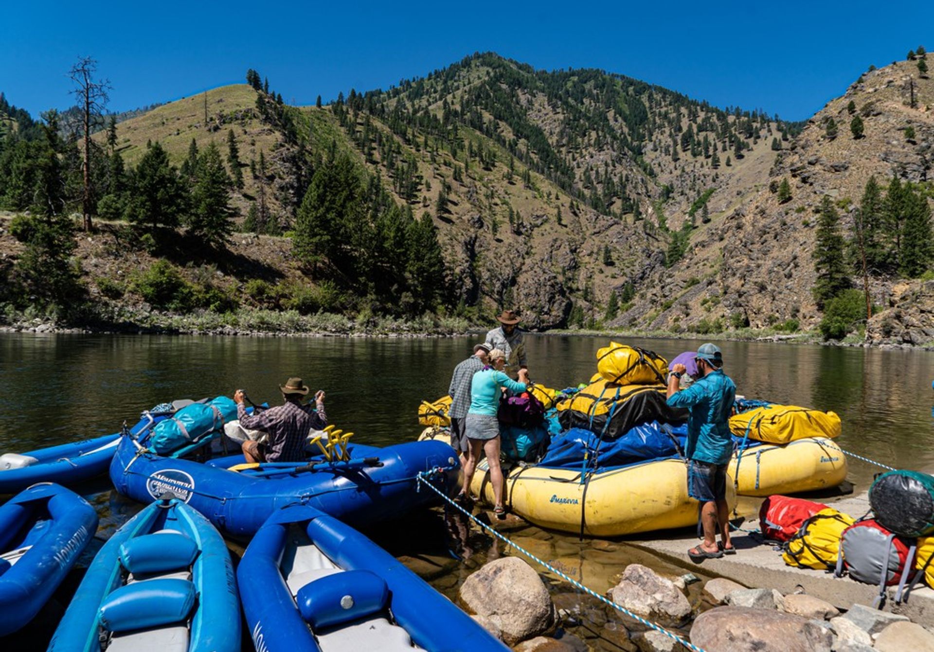 Group prepping colorful inflatable rafts and yellow dry bags on a calm mountain riverbank with pine-covered canyon slopes and a bright blue summer sky — river rafting adventure.
