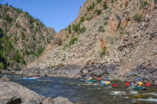 Four kayakers in inflatable kayaks wearing helmets and life jackets paddling with red paddles down a mountain river through a rocky canyon with steep cliffs, scattered pine trees and a clear blue sky.