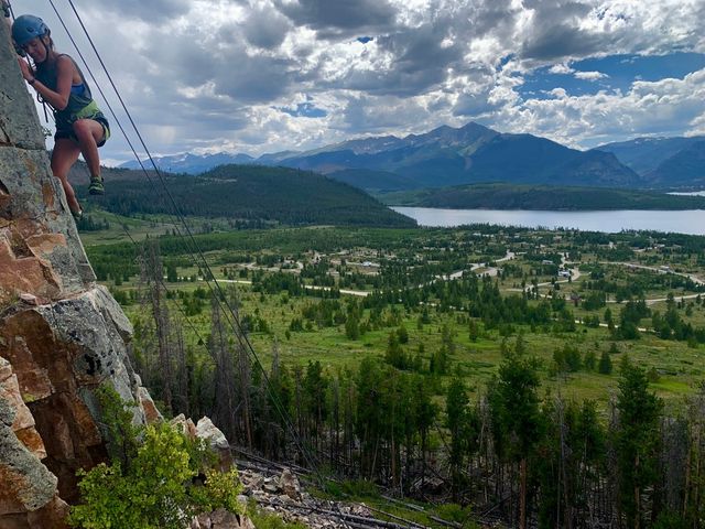 Adventurous rock climber in helmet and harness scaling a cliff above a green alpine valley and mountain lake beneath dramatic clouds.