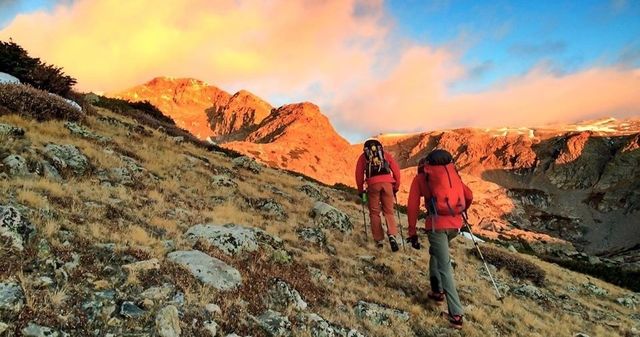 Two hikers with backpacks and trekking poles ascending a rocky alpine slope at sunrise, golden alpenglow illuminating rugged mountain peaks and grassy scree.