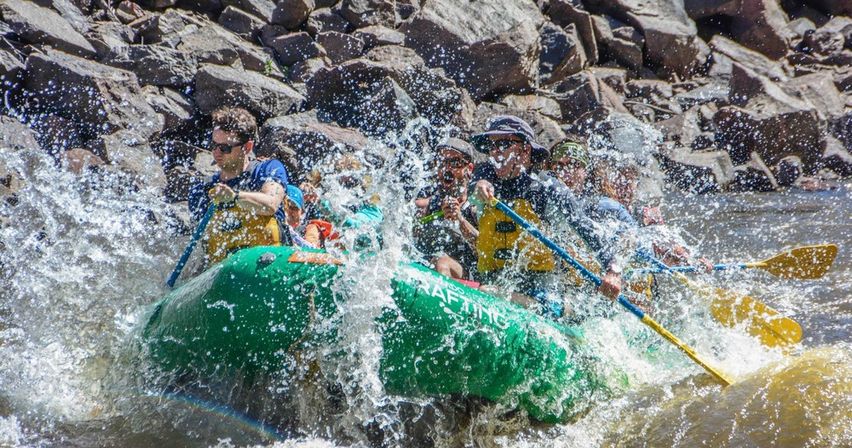 Whitewater rafting adventure with a group paddling a green inflatable raft through splashing river rapids beside rocky banks