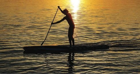 Silhouette of a stand-up paddleboarder gliding across calm golden water at sunset with the sun's reflection trailing behind.