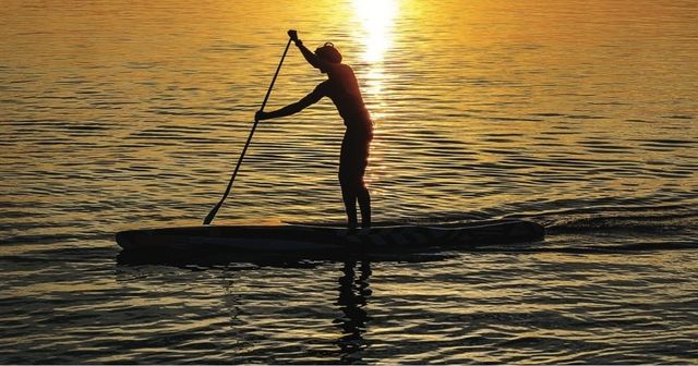 Silhouette of a stand-up paddleboarder gliding across calm golden water at sunset with the sun's reflection trailing behind.