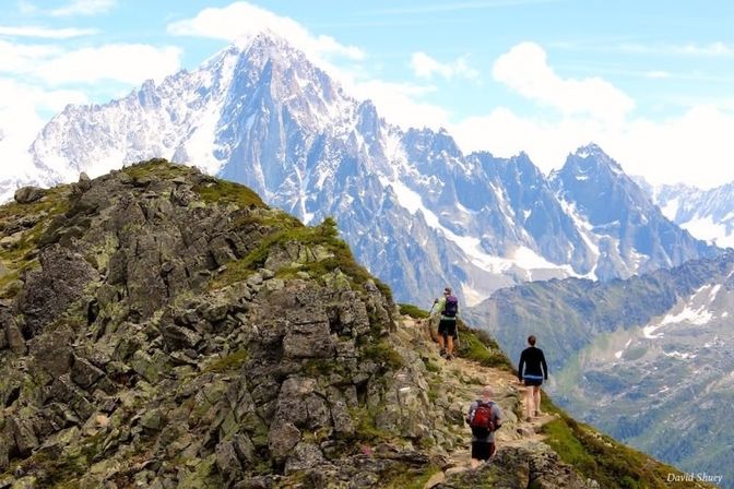 Three hikers with backpacks on a rocky alpine ridge trail heading toward towering snow‑capped peaks under a bright blue sky