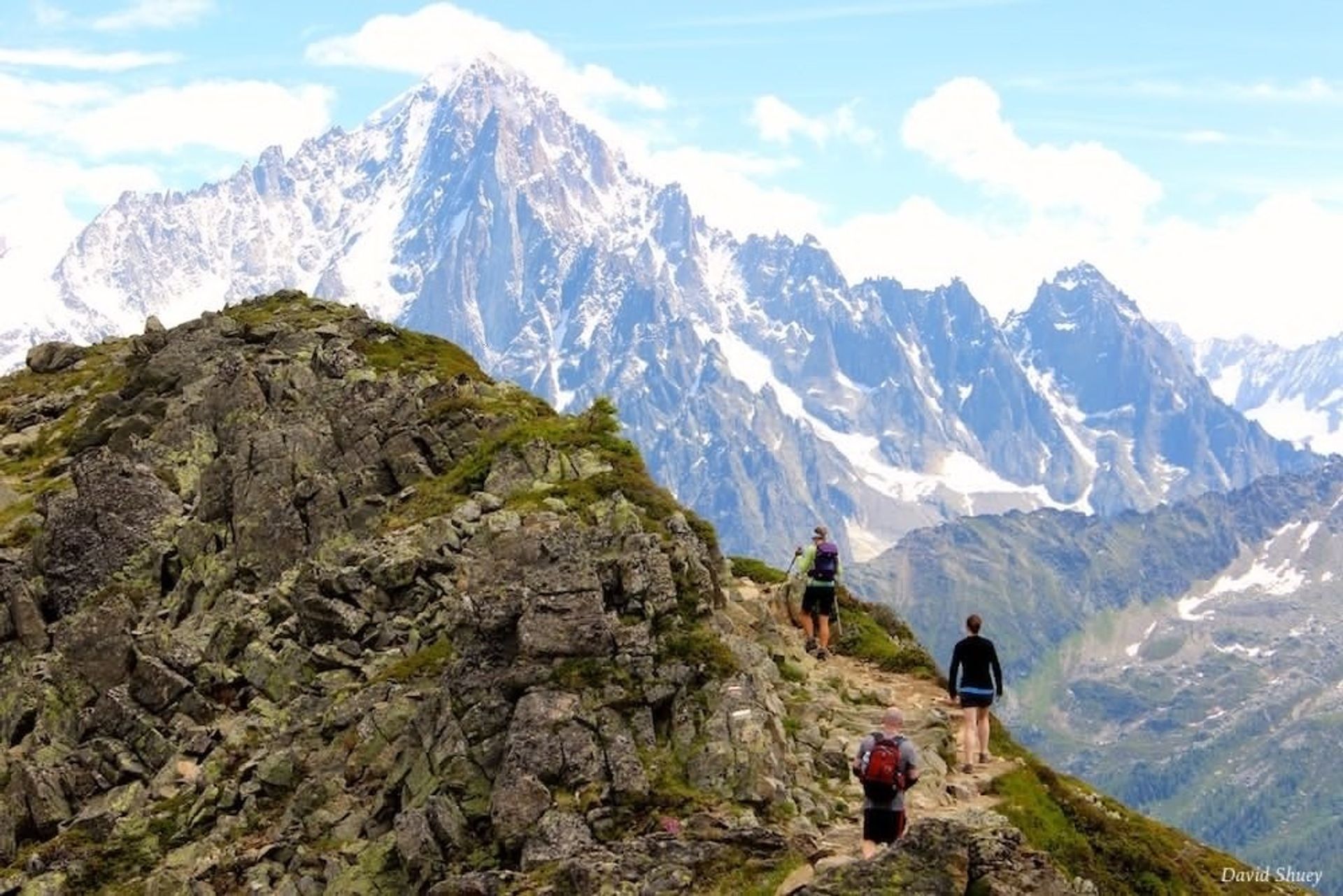 Three hikers with backpacks on a rocky alpine ridge trail heading toward towering snow‑capped peaks under a bright blue sky