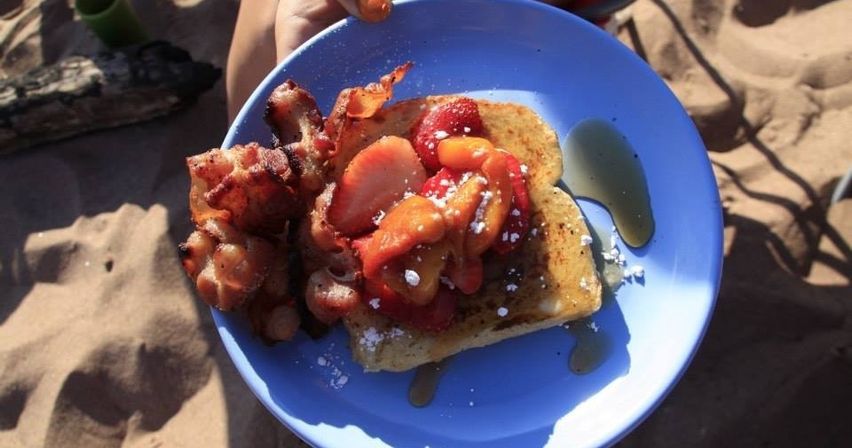 Beach breakfast: blue plate with French toast topped with sliced strawberries, syrup and powdered sugar, plus crispy bacon on a sandy beach