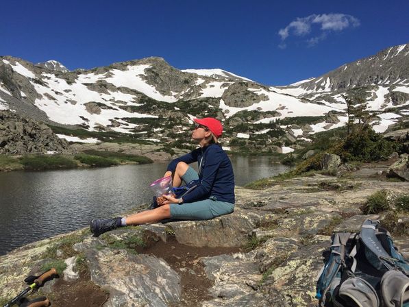 Hiker wearing a red cap and blue jacket sitting on a rocky shore eating a snack beside a backpack and trekking poles at an alpine lake, with snow-patched rugged mountain peaks and a clear blue sky in the background