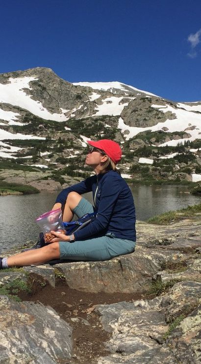 Hiker wearing a red cap and blue jacket sitting on a rocky shore eating a snack beside a backpack and trekking poles at an alpine lake, with snow-patched rugged mountain peaks and a clear blue sky in the background