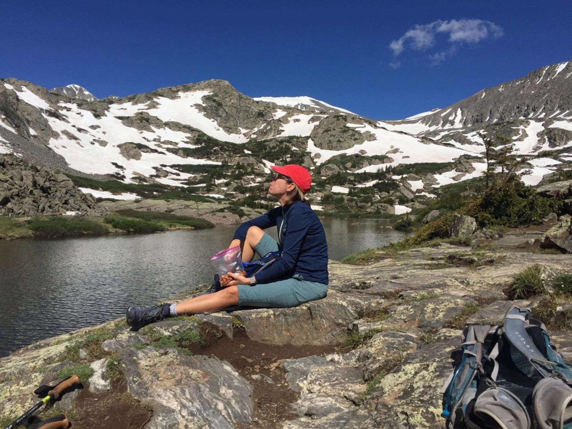 Hiker wearing a red cap and blue jacket sitting on a rocky shore eating a snack beside a backpack and trekking poles at an alpine lake, with snow-patched rugged mountain peaks and a clear blue sky in the background