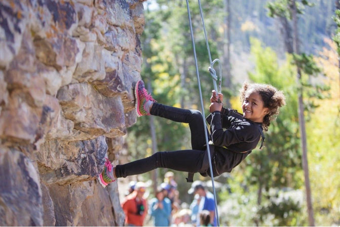 Smiling young girl rock climbing on a sunlit natural rock face, secured by rope and harness with forested trees and onlookers blurred in the background.