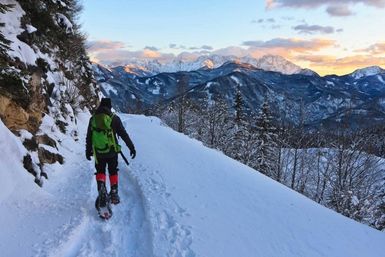 Person snowshoeing on a narrow snowy mountain trail at sunset with a bright green backpack and red gaiters, overlooking snow-covered alpine peaks and pine forest