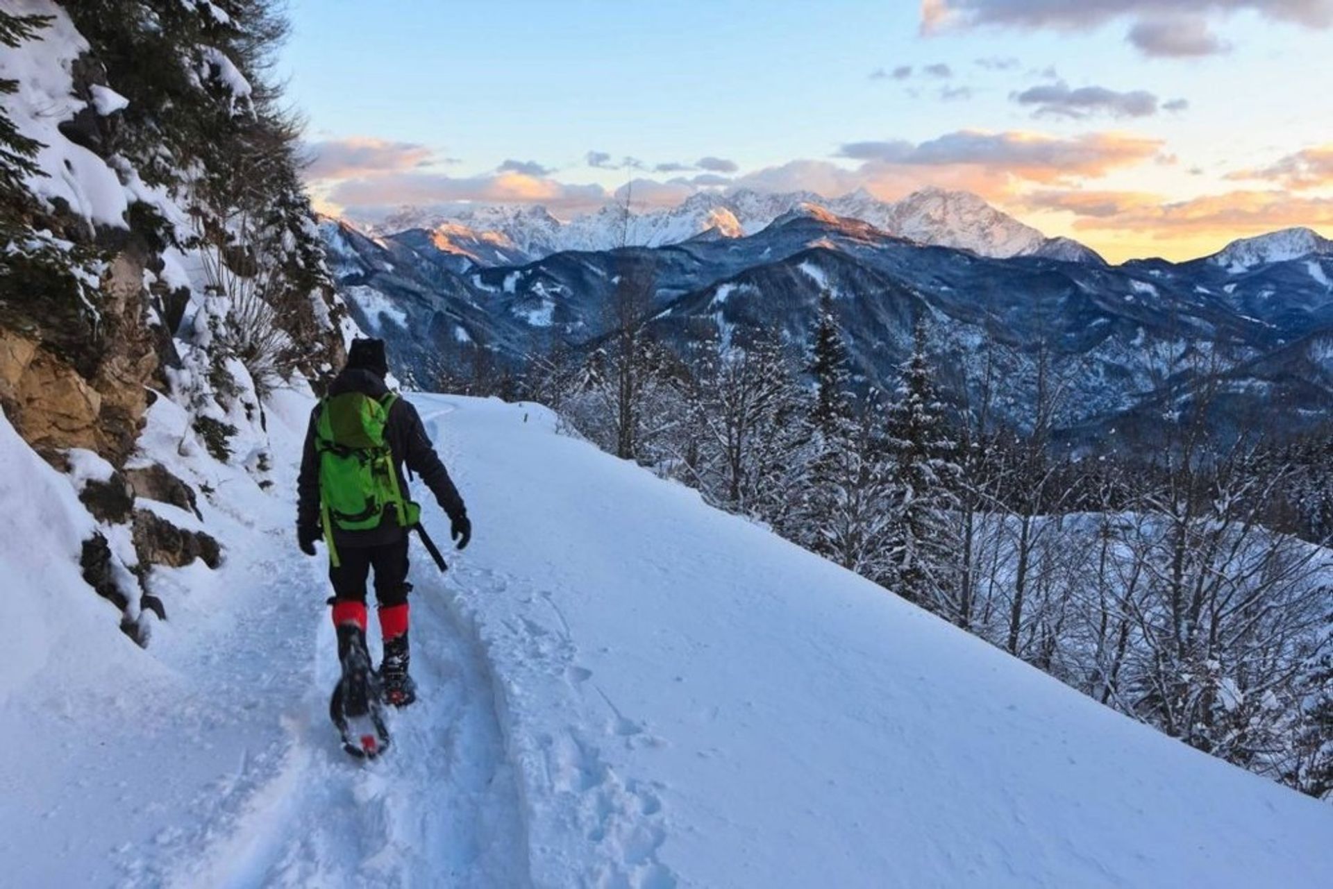 Person snowshoeing on a narrow snowy mountain trail at sunset with a bright green backpack and red gaiters, overlooking snow-covered alpine peaks and pine forest