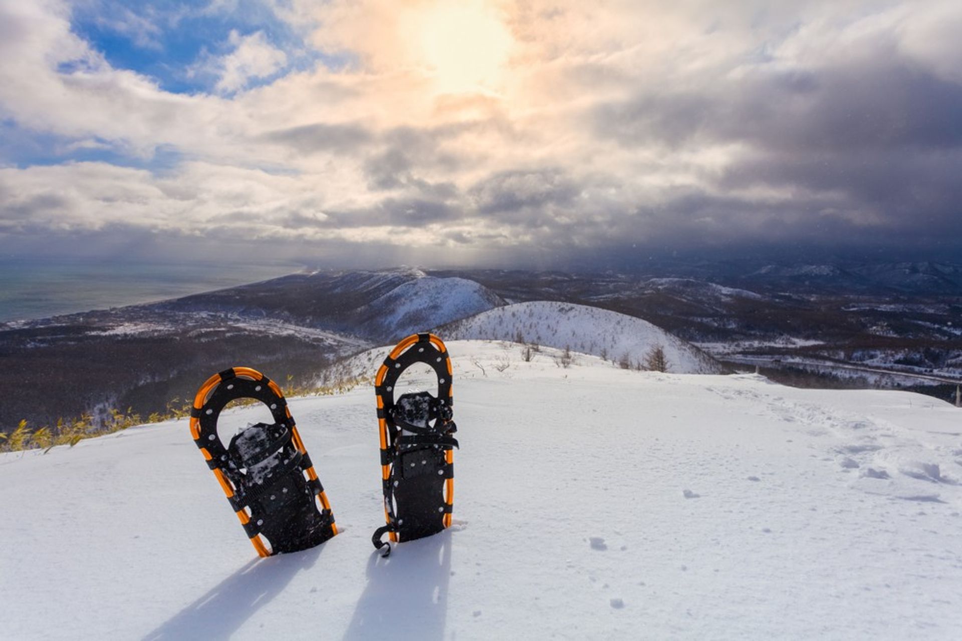 Orange snowshoes standing in fresh powder on a snowy mountain ridge, panoramic winter peaks under dramatic sunrise clouds