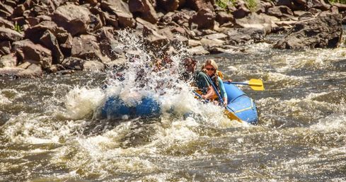 Thrilling whitewater rafting on a rocky mountain river — people in a blue inflatable raft hit churning rapids with a big splash and yellow paddle.