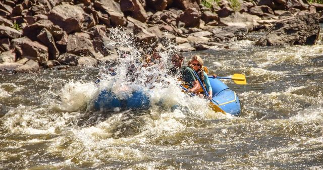 Thrilling whitewater rafting on a rocky mountain river — people in a blue inflatable raft hit churning rapids with a big splash and yellow paddle.