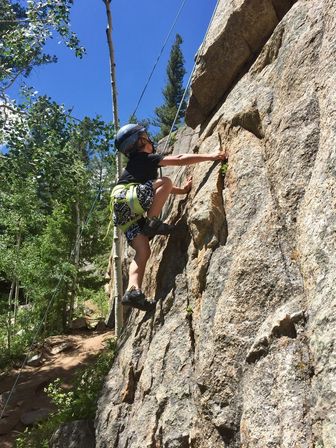 Child rock climber wearing a helmet and harness scales a granite cliff on a sunny day with a forested trail and blue sky in the background.